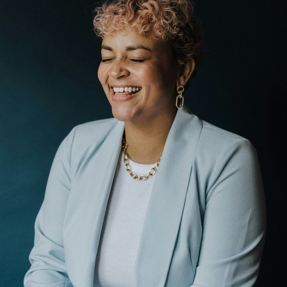 Woman in light blue blazer with gold necklace and earrings against a dark background