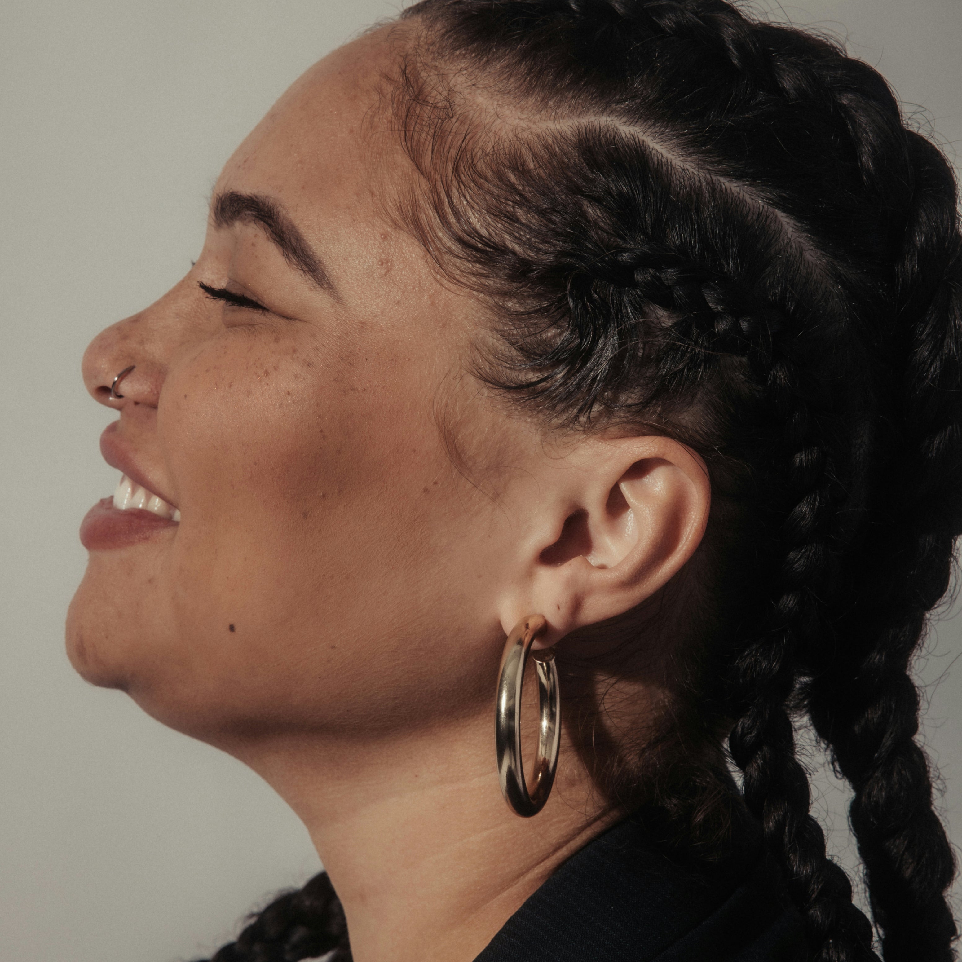 Side profile of a woman with braided hair wearing a gold hoop earring against a neutral background